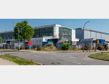 Industriegebäude mit großen Glasfenstern und weißer Fassade, umgeben von einem Zaun mit Stacheldraht. Im Vordergrund befinden sich gestapelte Paletten, Bagger und eine asphaltierte Straße mit einem Straßenschild.