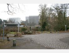 Pflasterweg in einem parkähnlichen, kahlen Gelände mit Büschen und Bäumen im Winter; im Hintergrund niedrige Gartenhäuser und mehrstöckige Wohnblocks.