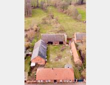 Luftaufnahme eines ländlichen Gehöfts mit mehreren Backstein- und Fachwerkgebäuden, die einen rechteckigen Innenhof umrahmen. Im Hintergrund sind grüne Wiesen, vereinzelt stehende Bäume und ein kleines Waldstück sichtbar.