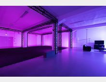 Ein moderner, offener Raum mit einer Konstruktion aus schwarzen Metallstreben, lila Beleuchtung und glattem, hellen Boden. Im rechten Bereich befinden sich eine schwarze Couch, ein Fernseher und ein kleiner Tisch, die einen Lounge-Bereich bilden.