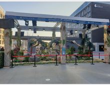 Eine Outdoor-Ausstellung mit einem Dinosaurier-ähnlichen Roboter-Modell, inspiriert von der Spieleserie „Horizon“, umgeben von künstlicher Vegetation und dekorativen Elementen. Im Hintergrund sind Schilder mit „Mercedes Platz“ und moderne Gebäude, darunter das Hotel Indigo, zu sehen.
