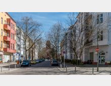 Straßenszene in einer Wohngegend mit mehrstöckigen, farbenfrohen Gebäuden auf beiden Seiten, parkenden Autos entlang der Straße und kahlen Bäumen. Im Hintergrund steht eine rote Backsteinkirche mit einem Kirchturm.