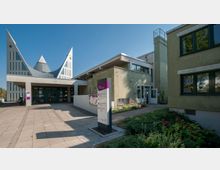 Moderner Kirchenkomplex mit markanter Architektur, darunter zwei spitze, nach oben geschwungene Dachformen. Rechts befinden sich angrenzende Gebäude mit grün verputzten Fassaden, davor ein kleiner bepflanzter Garten und ein Hinweisschild im Eingangsbereich.