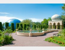 Ein Garten mit geometrischen Wasserbecken, umgeben von gepflegten Hecken und blühenden Blumen. Im Hintergrund befinden sich eine gläserne, blaue Kuppel sowie ein orientalisch anmutendes Pavillon mit Bögen und einer verzierten weißen Kuppel.