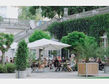 Ein Außenbereich eines Cafés oder Restaurants mit weißen Sonnenschirmen, runden Tischen und Stühlen, umgeben von Pflanzen in Holzkübeln und Bäumen. Im Hintergrund ist eine mit Efeu bewachsene Wand mit Fenstern sowie eine steinerne Treppe mit Balustrade zu sehen.