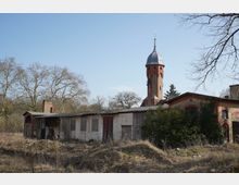 Blick auf ein Gelände mit niedrigen, teils verfallenen Backstein- und Putzgebäuden im Vordergrund; dahinter ragt ein hoher runder Backsteinturm mit Metallkuppel und Spitze auf. Umgeben ist die Anlage von trockenem Bewuchs und kahlen Bäumen unter klarem Himmel.