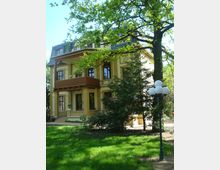 Zweistöckige Villa aus gelben Backsteinen mit Balkon und Walmdach, umgeben von grünen Bäumen und einer gepflegten Rasenfläche; im Vordergrund steht eine Straßenlaterne mit Kugelleuchten.