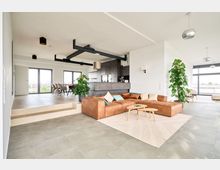 Modernes Wohnzimmer in offener Bauweise, mit einer großen braunen Ledercouch, einem hellen Teppich und einem kleinen Holztisch. Im Hintergrund eine Küche mit grauen Schränken, ein Essbereich mit Tisch und Stühlen sowie bodentiefe Fenster, die viel Tageslicht hereinlassen.