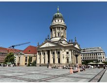 Neubarocke Kirche mit großem Kuppelbau und Säulenfassade, umgeben von einem weiten, gepflasterten Platz mit Sitzbänken und Menschen. Im Hintergrund moderne und historische Gebäude sowie ein Kran.