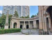 Steinerne, halbkreisförmige Kolonnaden mit verzierten Bögen und Reliefs, flankiert von einer Säule (Obelisk) auf der linken Seite und umgeben von grünen Bäumen; im Hintergrund ist ein modernes, hohes Wohngebäude zu sehen.