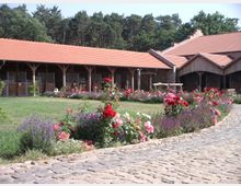 Ein gepflasterter Hof mit bunten Blumenbeeten und Lavendel führt zu einem langgestreckten Gebäude mit rotem Ziegeldach und offenen Stalltoren. Im Hintergrund gibt es einen Wald mit Nadelbäumen.