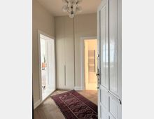 Offener Eingangsbereich einer Wohnung mit Parkettboden, einem roten Teppichläufer und einer modernen, weißen Einbauschrankwand. Links führt eine Tür in einen weiteren Raum, in dem ein kleiner Hund sichtbar ist, und rechts eine Tür zu einem Badezimmer mit Handtuchheizkörper.