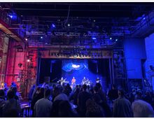 Ein Konzert in einem Industriestil-Veranstaltungsraum mit Backsteinwänden und sichtbaren Metallstrukturen. Auf der Bühne spielen Musiker vor einem beleuchteten Hintergrund mit galaktischem Motiv, während Zuschauer im Vordergrund stehen.