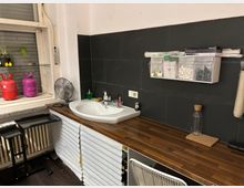 Kleiner Küchen- oder Arbeitsbereich mit langem Holzarbeitsplattenblock und eingelassenem Waschbecken vor einer dunklen Fliesenrückwand. Links befindet sich ein Fenster mit Ablage und Hockern, rechts hängen transparente Aufbewahrungsboxen und es stehen diverse Utensilien auf der Arbeitsfläche.