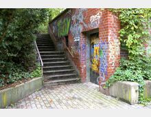 Backsteinbau mit Eingangstür, die mit Graffiti und Plakaten bedeckt ist, daneben eine Treppe mit Metallgeländer, die nach oben führt. Umgeben von dichter Vegetation und Pflastersteinen am Boden.