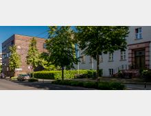 Straßenszene mit einem modernen Gebäude aus dunklem Backstein auf der linken Seite und einem hellen historischen Gebäude mit rosa-umrahmtem Eingang auf der rechten Seite. Die Szenerie ist von Bäumen und Hecken gesäumt, mit parkenden Autos im Vordergrund.