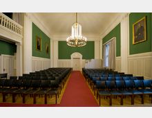 Innenansicht eines eleganten Versammlungsraums mit Stuhlreihen, grün gestrichenen Wänden, weißer Wandvertäfelung und großen, dekorativen Kronleuchtern. An den Wänden hängen gerahmte Porträts, und ein roter Teppich führt zu einer weißen Tür mit verzierter Umrahmung.