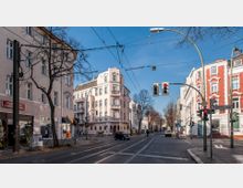 Straßenkreuzung in einem urbanen Wohnviertel mit mehrstöckigen Altbaugebäuden in hellen Pastell- und Rot-Tönen. Oberleitungen für Straßenbahnen, Ampeln und Straßenschilder prägen das Bild, während die Bäume am Straßenrand teilweise kahl sind.