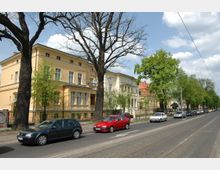 Straßenansicht mit einer Reihe historischer Wohnhäuser, darunter ein gelbes Gebäude im Vordergrund, flankiert von hohen Bäumen. Entlang der Fahrbahn mit Straßenbahngleisen parken mehrere Autos.