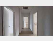 Ein leerer Flur in einem Büro- oder Wohngebäude mit weißen Wänden, mehreren offenen Türen zu angrenzenden Räumen und einer Deckenbeleuchtung mit Gittern. Der Boden besteht aus schlichtem, beigem Belag.