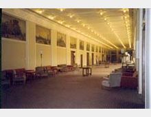 Langer, heller Innenraum mit einer Reihe von Wandgemälden auf beiden Seiten, einer hohen Decke mit einer Vielzahl von Lichtern und verstreuten Sitzgelegenheiten wie Sofas und Sesseln. Die Fenster befinden sich auf der rechten Seite des Raumes.