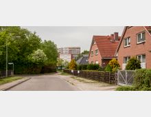 Straßenszene in einem Wohngebiet mit Einfamilienhäusern aus roten Ziegeln und roten Satteldächern. Im Hintergrund stehen Plattenbauwohnhäuser, und der Straßenrand ist von Bäumen und Hecken gesäumt.