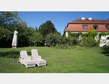 Ein weitläufiger Garten mit einer Rasenfläche, auf der zwei weiße Liegestühle und ein geschlossener Sonnenschirm stehen. Im Hintergrund befindet sich ein Gebäude mit rotem Ziegeldach und bewachsenen Fassaden, umgeben von Bäumen und Sträuchern.