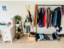 Ein Schlafzimmer oder Ankleidebereich mit einer weißen Kommode, einer hölzernen Pflanzentreppe mit Topfpflanzen, einem Kleiderständer mit Kleidung und Taschen sowie einem geflochtenen Korb auf dem Boden. Der Raum hat helle Wände und einen beigefarbenen Teppichboden.