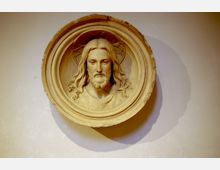 Rundes Relief aus Stein oder Gips mit einer Darstellung von Jesus Christus mit langem Haar und Bart, eingerahmt von einem dekorativen Kreis, vor einer hellen Wand.