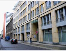 Ein moderner, mehrstöckiger Bürogebäudekomplex mit beiger Fassade und großen Fenstern entlang einer städtischen Straße. Im Vordergrund parken Autos, und ein Fußgänger steht nahe einem Eingang mit Glasvordach.