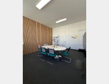 Moderner Besprechungsraum mit einem weißen, runden Tisch, umgeben von sechs gepolsterten Stühlen mit Metallrahmen. Eine Wand ist mit vertikalen Holzpaneelen verkleidet, die andere ist weiß und zeigt ein Logo mit der Aufschrift „Gexx aero sol“. Im Hintergrund steht ein Regal mit Broschüren, und die Decke ist mit rechteckigen Deckenleuchten ausgestattet.