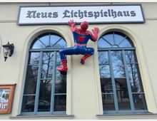 Fassade eines Gebäudes mit der Aufschrift „Neues Lichtspielhaus“, zwei großen Bögenfenstern und einer angebrachten Spiderman-Figur, die scheinbar an der Wand klettert. Links befindet sich eine Laterne und ein Schaukasten mit Veranstaltungsplakaten.