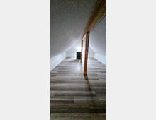 Ein länglicher Dachbodenraum mit niedriger Decke und schrägen Wänden, Holzstützbalken in der Mitte und einem kleinen Fenster am Ende. Der Boden ist mit rustikalen Laminatdielen ausgelegt.