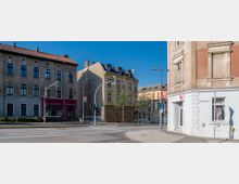 Straßenecke mit historischen Gebäuden, darunter ein mehrgeschossiges Haus mit rotbraunem Dach und ein hellgelbes Gebäude mit verzierten Fassadenelementen. Im Vordergrund eine Ampelkreuzung mit Gehweg und Straßenlaternen.