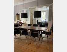 Großzügiger Raum mit einem Holztisch und sechs schwarzen Drehstühlen auf einem Holzboden, beleuchtet von zwei hängenden schwarzen Lampenschirmen. Im Hintergrund sind geöffnete Flügeltüren, die in ein helles Wohnzimmer führen, sichtbar, mit großen Fenstern und moderner Einrichtung.