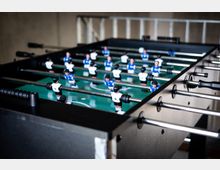 Ein Tischfußballspiel mit einem Spielfeld in Grün, Spielern in blauer und weißer Kleidung, Metallstangen und schwarzen Griffen steht in einem Innenraum mit neutralem Hintergrund.