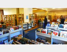 Innenansicht einer öffentlichen Bibliothek mit Bücherregalen, die mit Büchern und Zeitschriften gefüllt sind. Im Vordergrund stehen Regale mit Kinder- und Bilderbüchern, während im Hintergrund Personen an einem Informationsschalter und an Tischen arbeiten.