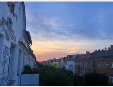 Straßenszene mit mehrstöckigen Wohnhäusern im Abendlicht. Links ist eine Fassade mit Stuckverzierungen und Balkonen zu sehen, im Hintergrund ein bunt gefärbter Himmel bei Sonnenuntergang.