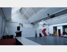 Ein moderner, heller Innenraum mit hohen Decken und sichtbaren Dachträgern. Der Raum verfügt über eine Bühne mit einem Rednerpult, Sitzgelegenheiten, eine weiße Wand mit Wandmalereien, eine offene Treppe aus Holz und Beleuchtungseinrichtungen an der Decke.