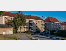 Mehrfamilienhäuser mit Putzfassaden und roten Ziegeldächern in einer Wohnsiedlung, umgeben von Grünflächen und Gehwegen; im Vordergrund ein gepflasterter Weg, parkende Autos und eine Straßenlaterne.