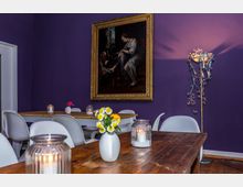 Ein gemütlicher Raum mit lila Wandfarbe, geschmückt mit einem großen Gemälde in einem goldenen Rahmen. Im Vordergrund stehen Holztische mit Dekorationen wie kleinen Vasen mit Blumen und Kerzenhaltern; im Hintergrund sind weiße Stühle und ein verschnörkelter Stehlichtständer sichtbar.