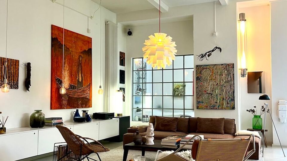 Open-plan modern living room with white walls and exposed ceiling beams, featuring a large grid-paned window and contemporary pendant lighting. The space includes wall art, low built-in cabinets, a brown sofa with lounge chairs on a large rug, and minimal decor on a polished light floor.