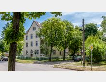 Mehrstöckiges Wohngebäude mit gelber Fassade, umgeben von Bäumen und einem weißen Zaun. Im Vordergrund eine gepflasterte Straße mit Verkehrsschildern und geparkten Autos in einer ruhigen Wohngegend.