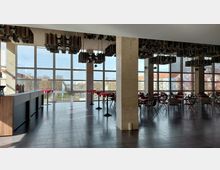 Großer moderner Veranstaltungsraum mit großen Glasfenstern, die viel Tageslicht hereinlassen. Im Raum stehen mehrere Tische und Stühle, dekoriert mit roten Tischdecken, während die Decke mit auffälligen, hängenden Metallleuchten gestaltet ist.