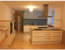 Moderne Küche mit hellen Holzböden, weißen Einbauschränken und einer zentralen Kochinsel mit Herdplatten. An der Rückwand befindet sich eine blaue Wand, ein Edelstahlwaschbecken, mehrere Steckdosen und ein Fenster, das natürliches Licht hereinlässt.