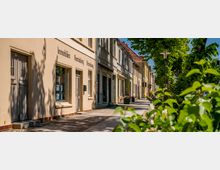 Straßenzug mit niedrigen, aneinandergebauten Altbaufassaden und Schaufenstern entlang eines breiten Gehwegs. Im Vordergrund rahmen grüne Bäume und Sträucher die Szene, daneben stehen Straßenlaterne und Ampel an der ruhigen Straße.