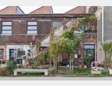 Backsteinindustriebau mit zweigeschossiger Fassade, teils verputzt und bewachsen. Im Vordergrund befinden sich ein kleiner Garten mit Bäumen und Blumen sowie eine graue Betonbank; ein schmaler Zugang führt zu einer Eingangstür mit der Aufschrift "Fundus".
