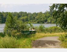 Zwei Personen sitzen auf einer grünen Bank in einem Park mit Blick auf einen Fluss, der von dichter Vegetation und Bäumen umgeben ist. Im Hintergrund ist ein kleiner Steg am gegenüberliegenden Ufer zu sehen.