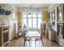 Wohnraum mit einem Holzboden und einer Mischung aus rustikalen und modernen Elementen, darunter eine freigelegte, teilweise verputzte Wand mit Bilderrahmen, ein schlichter Tisch mit bunten Stühlen und Regale mit Ordnern und Dekorationen. Große Fenster mit weißem Holzrahmen lassen viel Licht herein.