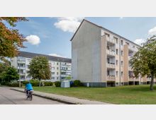 Mehrstöckige Wohngebäude mit hellen Fassaden und Balkonen, umgeben von Rasenflächen, Bäumen und Gehwegen. Eine Person fährt mit dem Fahrrad auf einem Weg, während drei Menschen auf einer Wiese im Hintergrund stehen.